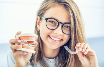 A smiling girl holdin a dental model with traditional braces in one hand and a clear aligner in the other.