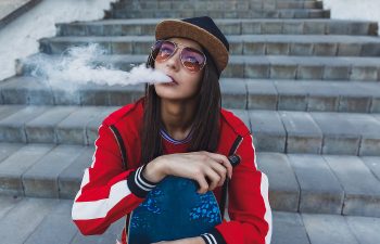 A person in a red jacket and sunglasses sits on steps, exhaling vapor from an e-cigarette, with a skateboard resting on their lap.