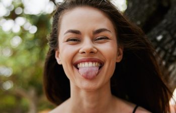 A young woman with long brown hair smiles playfully at the camera, sticking out her tongue. She is outdoors with blurred greenery in the background and sunlight on her face.